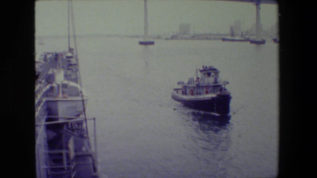 Small tugboat on calm harbor water seen from a shipboard vantage point, with railings in the foreground and a distant bridge or shoreline span in the background.