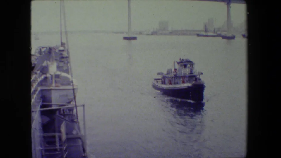 Small tugboat on calm harbor water seen from a shipboard vantage point, with railings in the foreground and a distant bridge or shoreline span in the background.