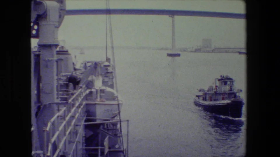 Tugboat crossing open harbor water from a shipboard perspective, with deck equipment and railings at left and a distant waterfront line under a hazy sky.