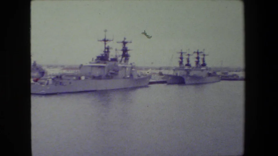 Gray naval vessels appear moored across calm harbor water, with tall mast structures and low shoreline buildings visible through faded 8mm color.