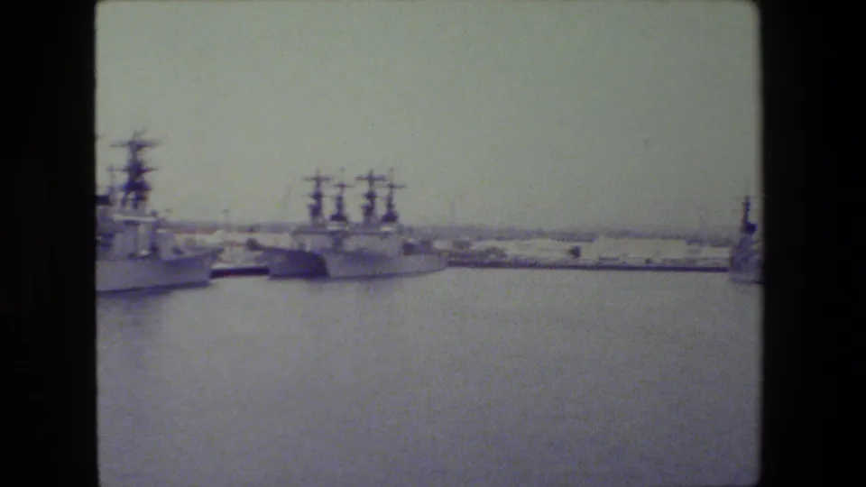 Docked gray naval ships line the far side of a calm harbor, with mast structures, shoreline infrastructure, and soft 8mm exposure across the frame.