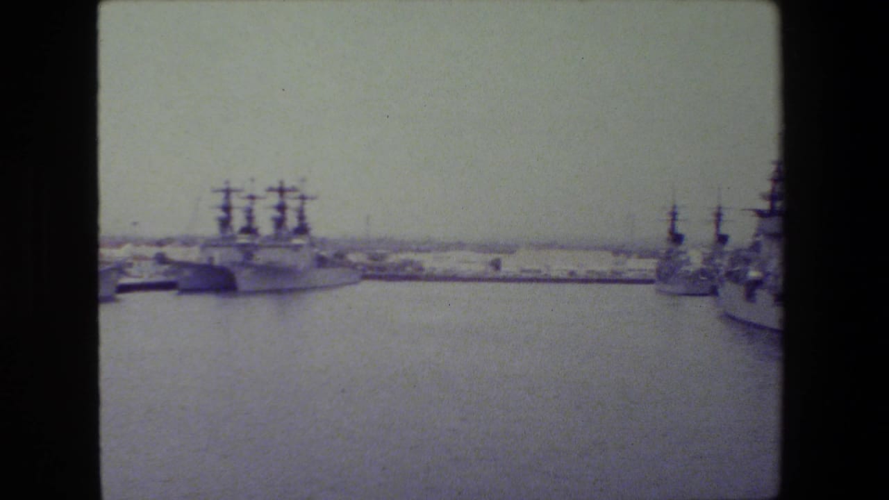 Soft 8mm frame showing a wide harbor view with gray naval vessels docked along both sides of the water under an overcast sky.