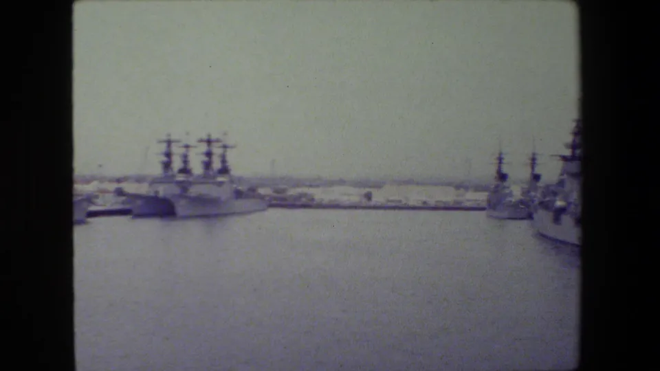 Soft 8mm frame showing a wide harbor view with gray naval vessels docked along both sides of the water under an overcast sky.