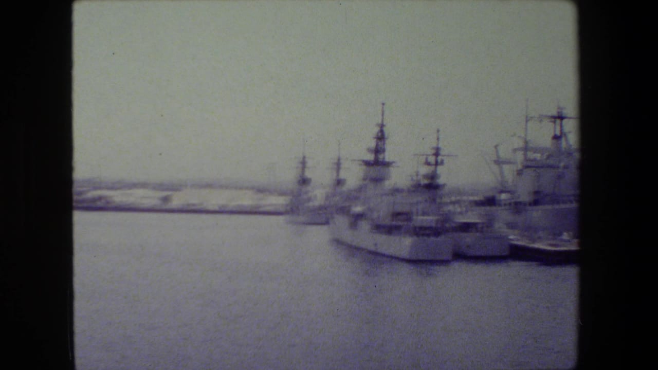 Naval ships docked along the right side of a harbor channel.