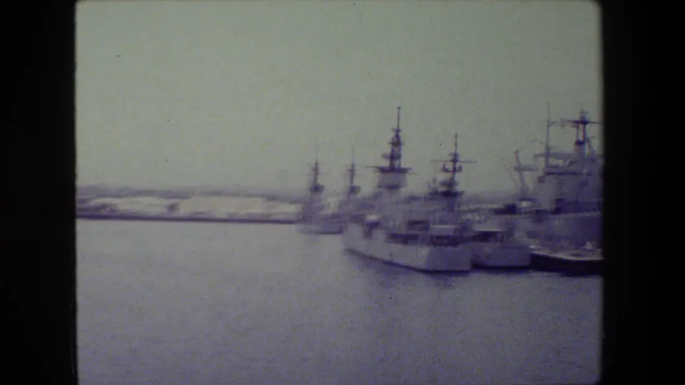 Panoramic 8mm view of multiple docked warships along a harbor edge, with additional vessels visible across the water.