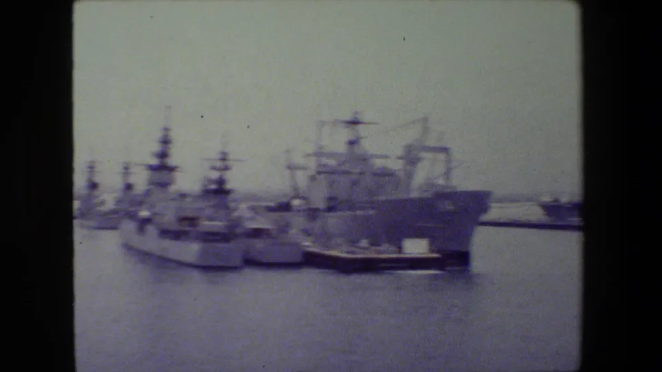 Soft-focus 8mm frame showing a large gray vessel near the center with smaller naval ships docked nearby in calm harbor water.