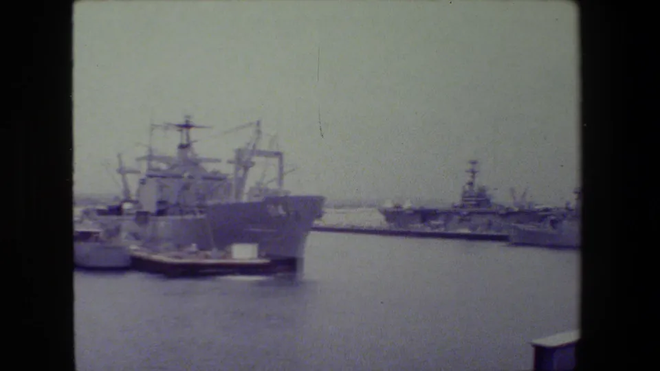 Final 8mm frame of the pan showing a large gray naval vessel in the foreground area with smaller warships and harbor structures in the distance.