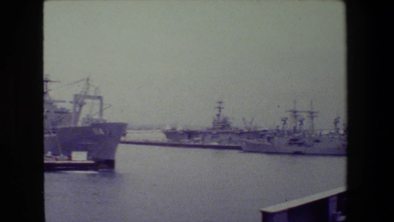 Distant U.S. Navy vessels at San Diego Naval Base in 1981, filmed across calm harbor water on faded 8mm stock.