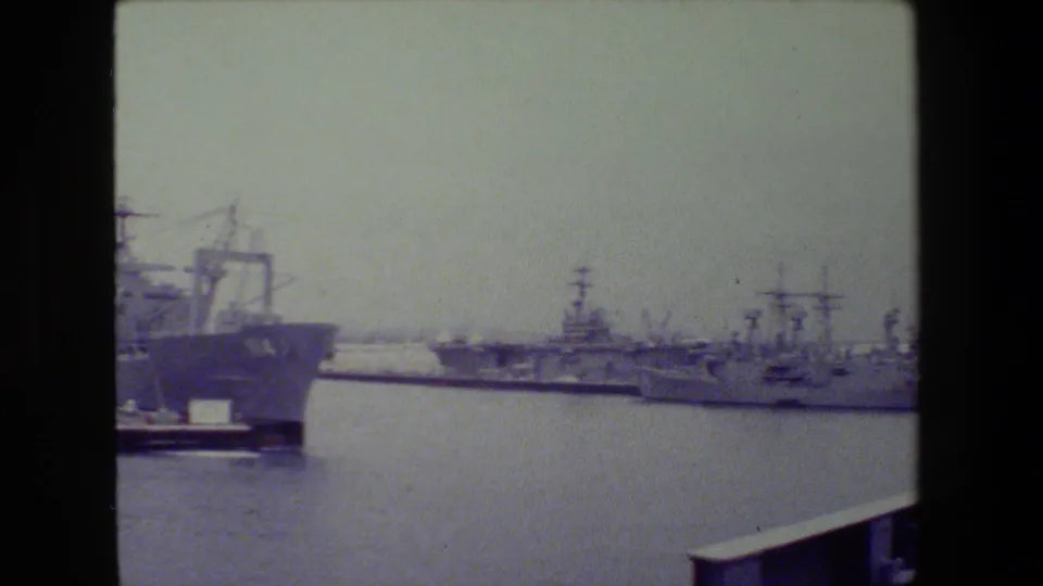 Distant U.S. Navy vessels at San Diego Naval Base in 1981, filmed across calm harbor water on faded 8mm stock.
