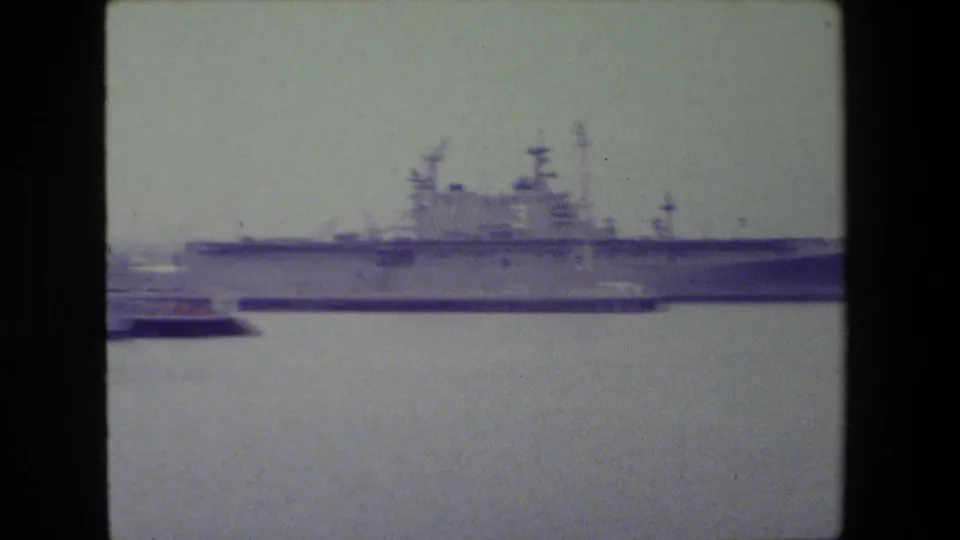 A large gray naval vessel, possibly with a broad flight deck or carrier-like profile, sits docked across the water in the 1981 San Diego naval harbor footage.