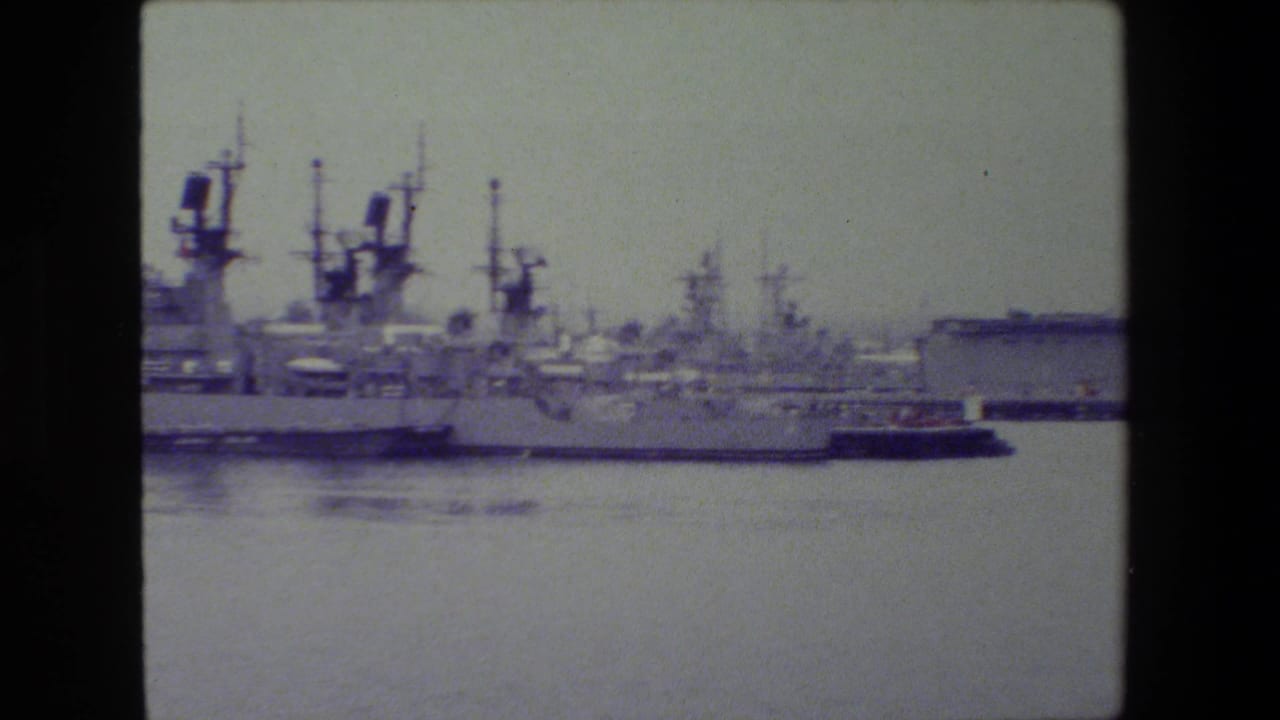 Soft 8mm view of multiple gray naval ships moored across calm water, with superstructures and antenna masts under a pale overcast sky.
