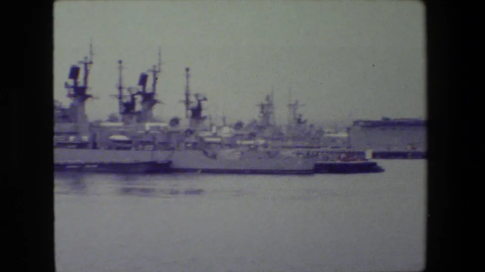 Soft 8mm view of multiple gray naval ships moored across calm water, with superstructures and antenna masts under a pale overcast sky.
