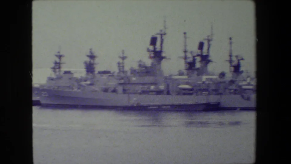 Harbor view of a large gray warship moored near other naval vessels, with radar masts and muted 8mm color visible across the frame.