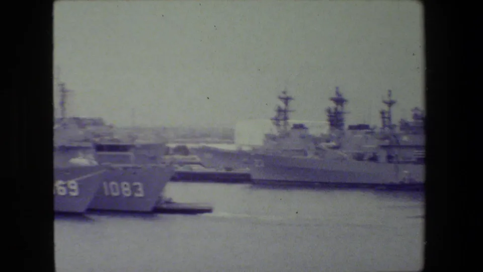 Handheld 8mm frame showing partial ship bows with visible hull numbers at left and larger docked naval ships across the water.