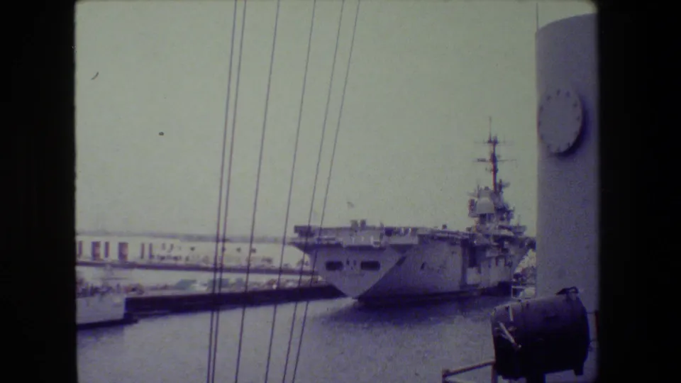 Soft 8mm harbor frame with an aircraft carrier-like vessel moored near a pier, vertical rigging lines in the foreground, and another ship structure at the right edge.