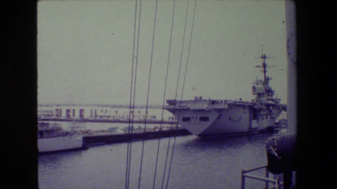 A gray naval vessel sits near a pier in archival 8mm footage, with vertical rigging or cables crossing the frame and another small craft or dock structure in the foreground.