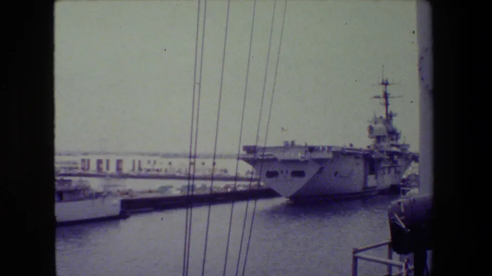 A gray naval vessel sits near a pier in archival 8mm footage, with vertical rigging or cables crossing the frame and another small craft or dock structure in the foreground.