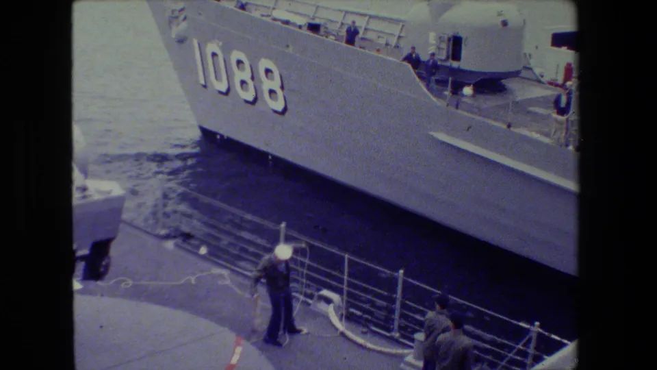 A large gray naval ship with hull number 1088 passes or rests close to a pier where sailors and mooring lines are visible below.
