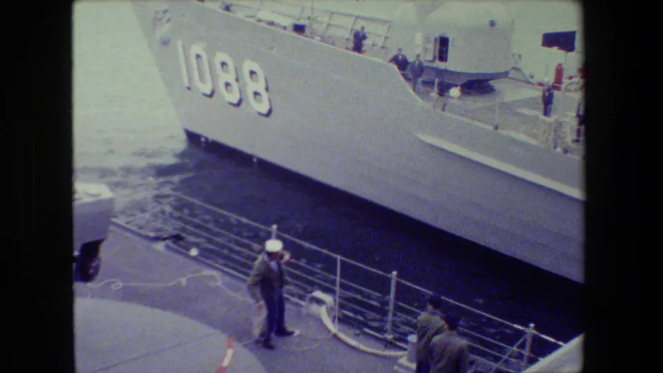 A sailor in a white cap stands near pier railings and rope while the gray naval ship marked 1088 occupies most of the frame.