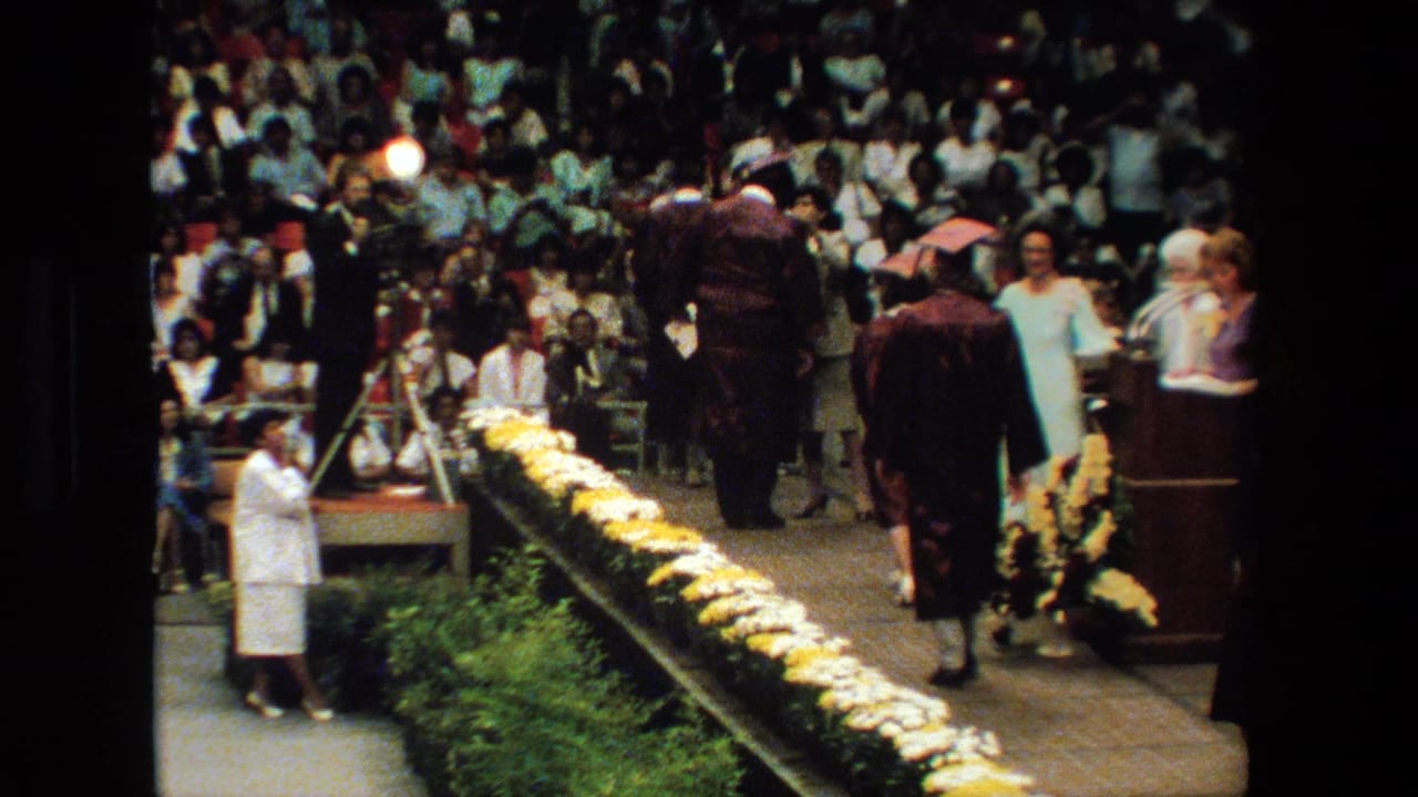 At a 1983 graduation ceremony in Lansing, Michigan, graduates in maroon caps and gowns move across a flower-lined stage while spectators watch from bleachers.