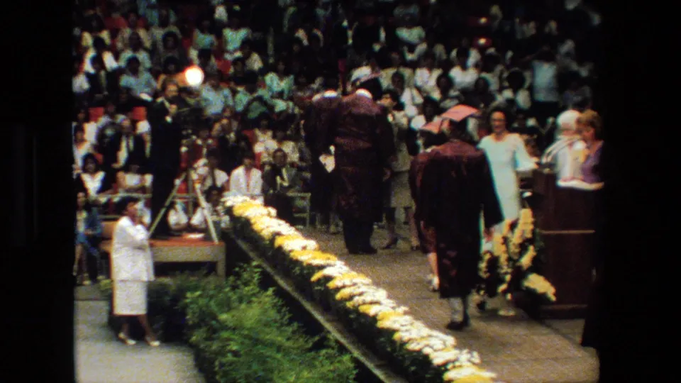 At a 1983 graduation ceremony in Lansing, Michigan, graduates in maroon caps and gowns move across a flower-lined stage while spectators watch from bleachers.