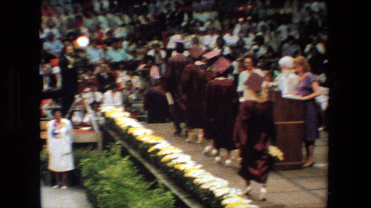 A graduate walks past ceremony staff on a stage with flowers and a seated crowd behind.