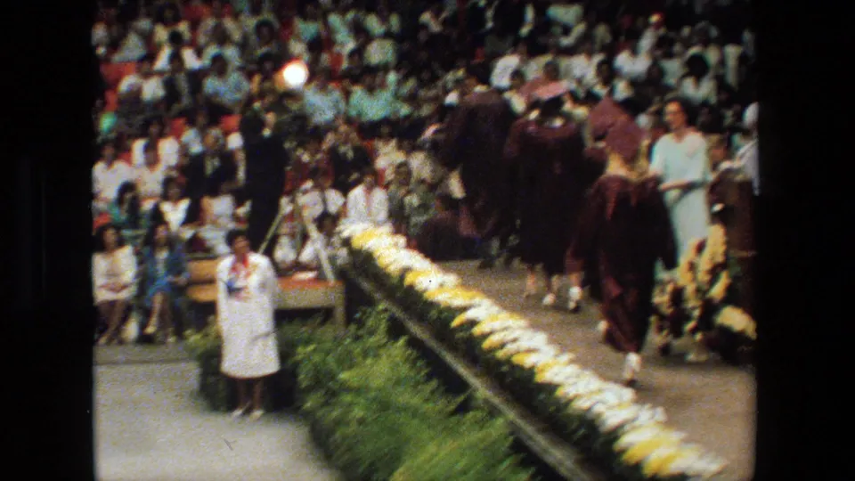 Multiple graduates in maroon gowns continue across the flower-lined stage at the 1983 Lansing graduation while a large audience watches from the bleachers.