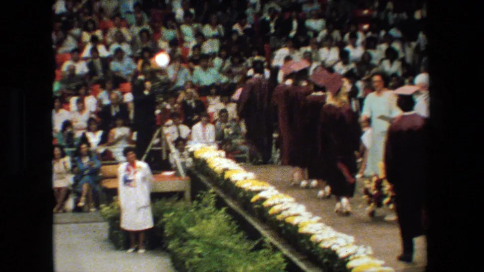 Graduates in maroon caps and gowns move away along the decorated stage during the 1983 Lansing, Michigan ceremony, framed by flowers and a crowded gym or auditorium setting.