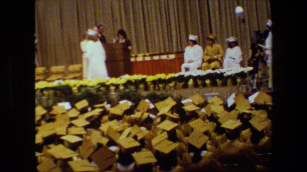 In this 1983 Lansing, Michigan graduation footage, rows of yellow mortarboards fill the foreground while people in white academic regalia and a speaker stand on a curtained stage.