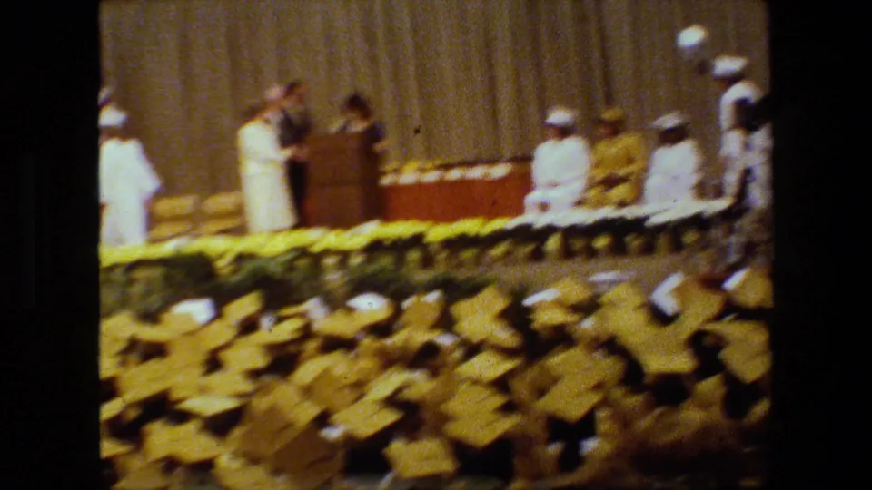 The ceremony continues from an audience angle, with a podium speaker on the curtained stage and yellow-capped graduates visible in the foreground.