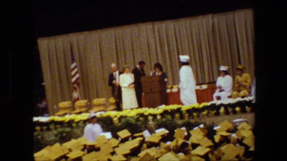 A stage presentation appears underway as adults stand near the podium, with yellow-capped graduates below and seated participants along the curtained backdrop.