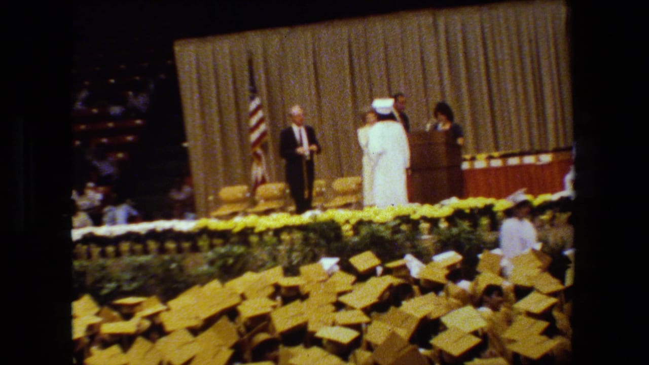 1983 Lansing, Michigan graduation ceremony with graduates in white caps and gowns near a stage, an American flag beside the curtain, and many yellow caps visible in front.