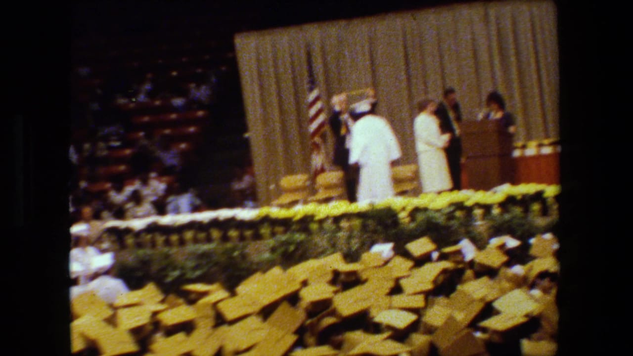 A graduate in white stands on the decorated stage during the Lansing graduation ceremony, with a suited official, American flag, beige curtain, and audience seating visible.