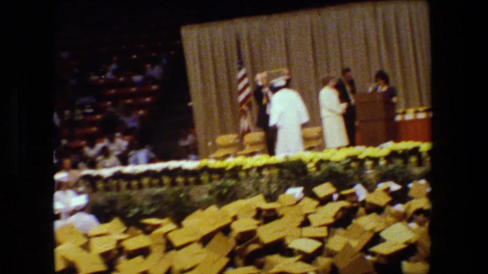 A graduate in white stands on the decorated stage during the Lansing graduation ceremony, with a suited official, American flag, beige curtain, and audience seating visible.