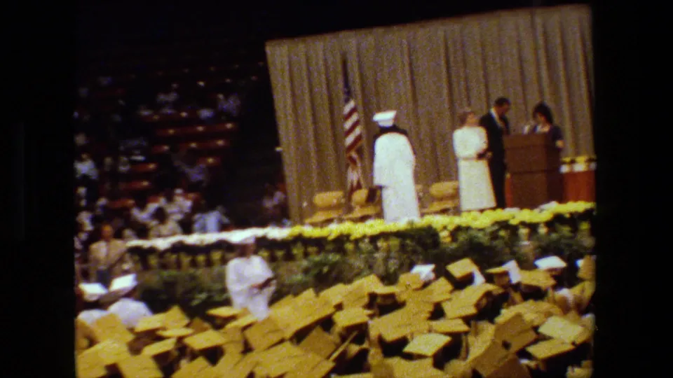 A white-gowned graduate pauses on the commencement stage in Lansing, Michigan, with a suited figure, podium, American flag, curtain backdrop, and yellow caps in the foreground.