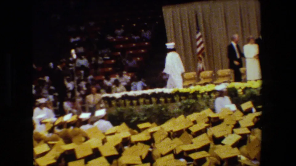 A graduate in a white cap and gown moves across the decorated graduation stage, with two adults near the beige curtain, the American flag, and rows of yellow caps below.