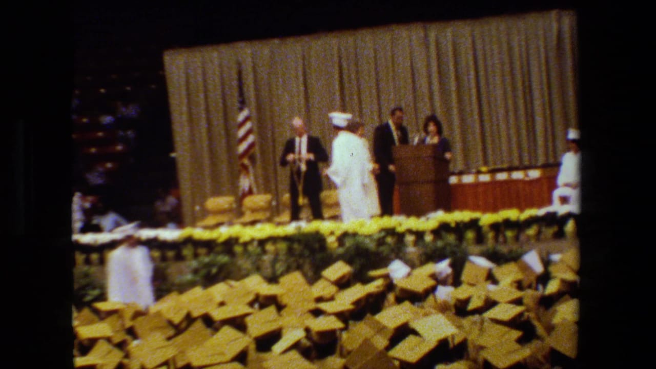 1983 Lansing graduation ceremony footage showing white-gowned graduates near a stage with officials, a podium, yellow flowers, and an American flag.