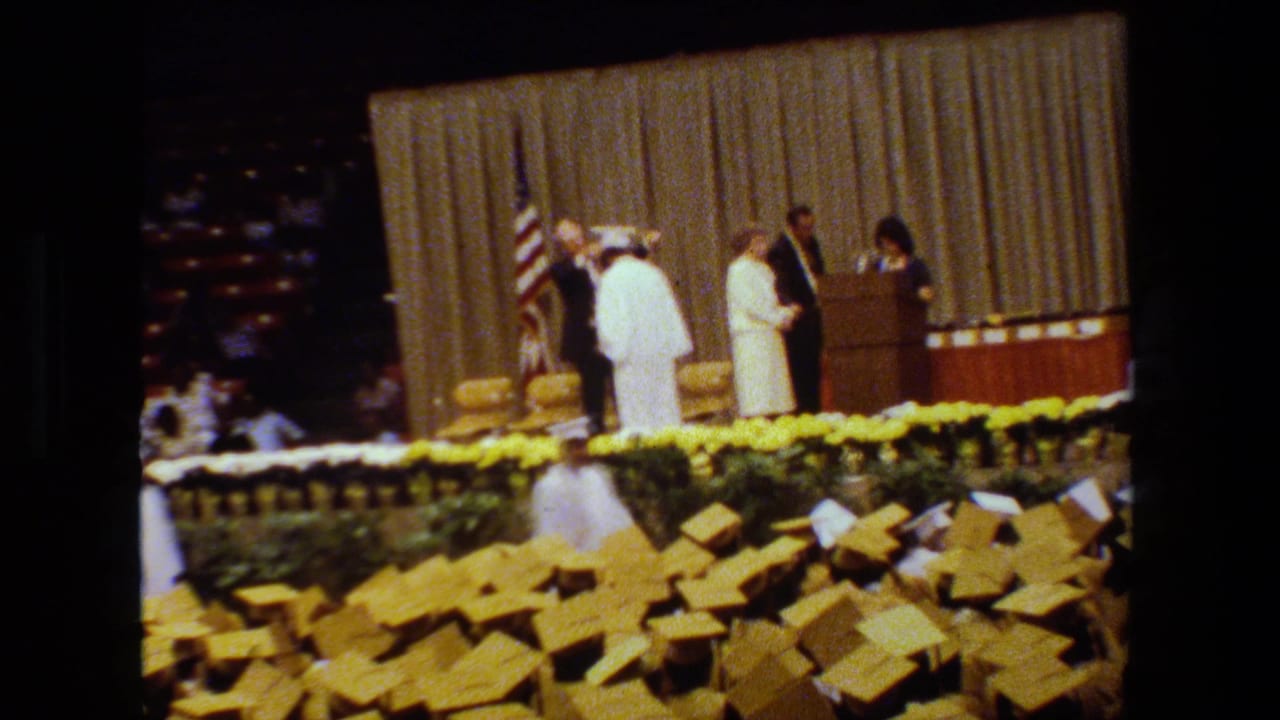 A graduate stands at the podium area as officials gather on a flower-lined stage.