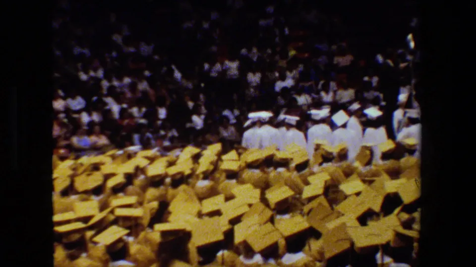 Audience-view 8mm frame of a 1983 Lansing graduation ceremony with many yellow mortarboards in the foreground and graduates in white near the stage.