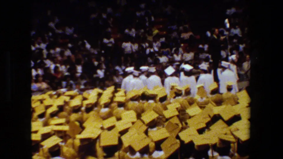 Wide audience-perspective frame from a 1983 Lansing graduation ceremony, showing graduates near the stage and many yellow caps across the foreground.