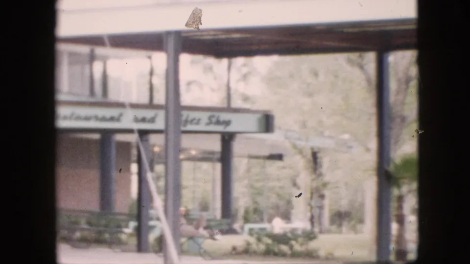 1958 Pennsylvania home-movie frame showing a low restaurant and gift shop building with visible signage, trees, outdoor seating, and a small landscaped area.