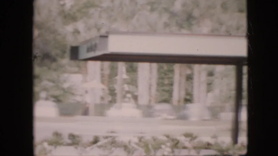 1958 Pennsylvania 8mm frame with a covered walkway or entrance structure crossing the view, seen through dark interior window framing above foreground shrubs.