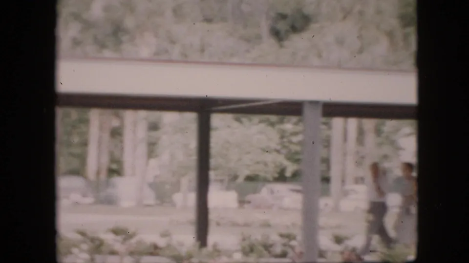 1958 Pennsylvania 8mm frame showing a covered structure, vertical posts, trees, shrubs, and two distant people standing near the right edge of the scene.