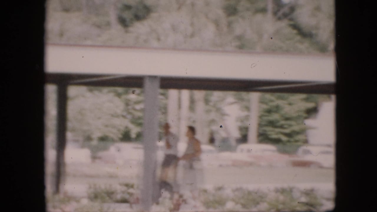 1958 Pennsylvania campus exterior viewed through a dark window or doorway, with a covered walkway, pillars, soft greenery, distant pedestrians, and parked cars.