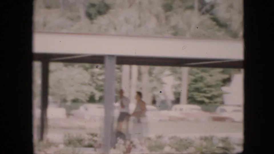1958 Pennsylvania campus exterior viewed through a dark window or doorway, with a covered walkway, pillars, soft greenery, distant pedestrians, and parked cars.