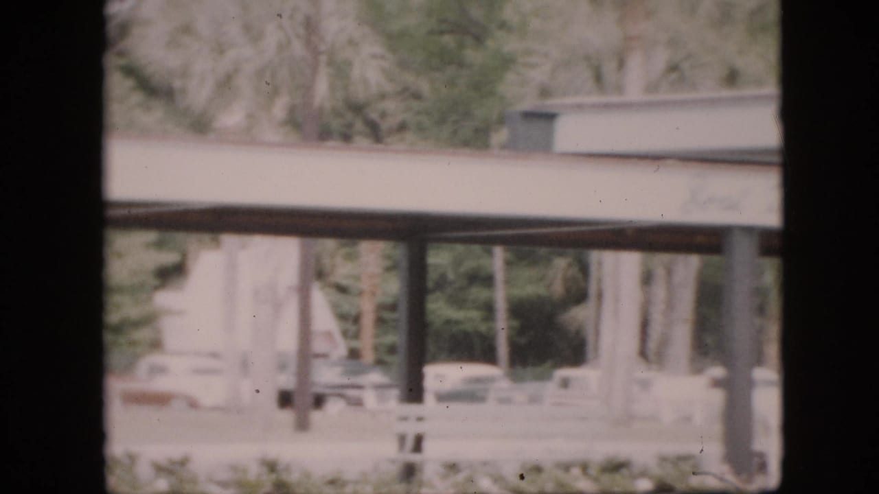 Covered walkway with pillars, trees, parked cars, and distant people viewed through a dark frame.