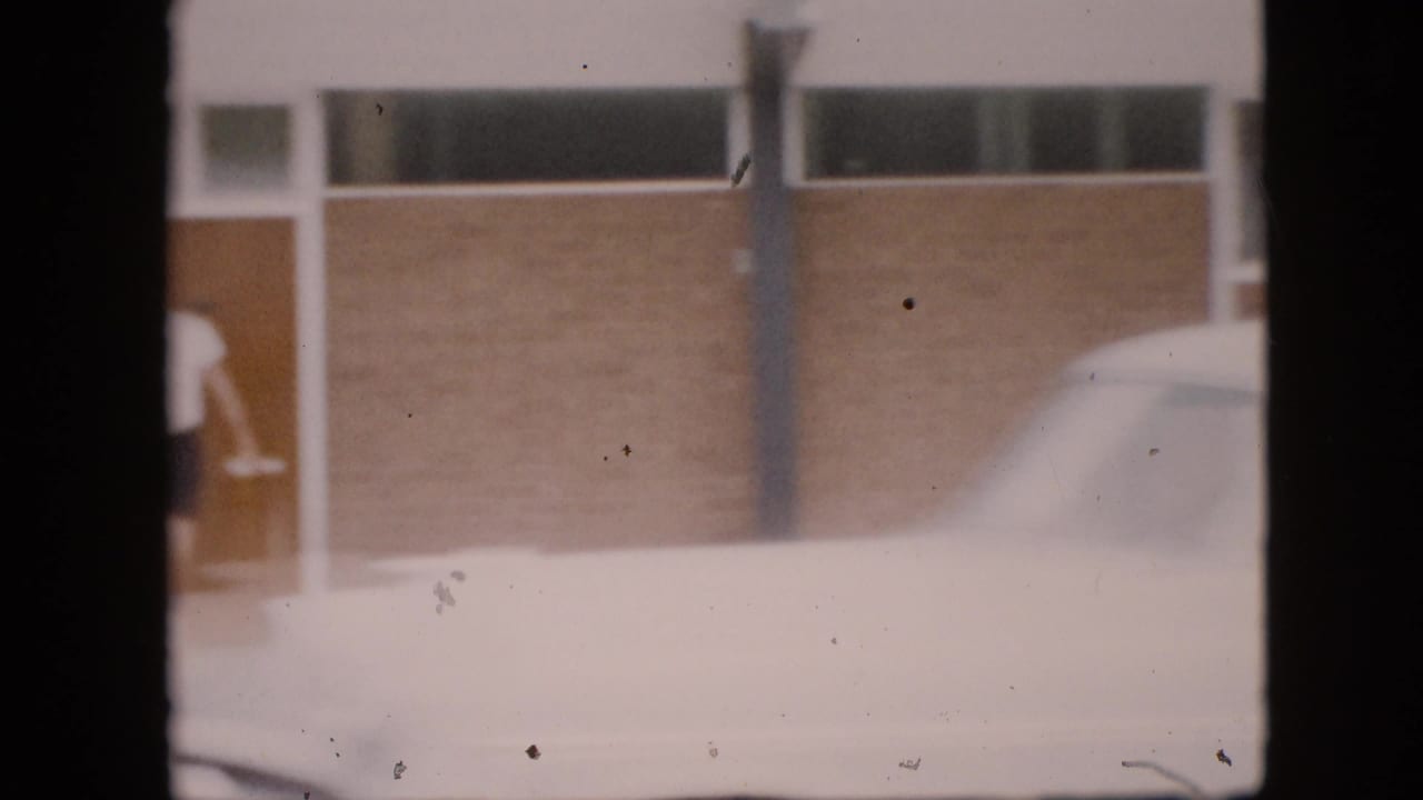 In 1958 Pennsylvania home-movie footage, a partial man in a white shirt and dark shorts stands near a glass-panel doorway beside a brick building and a white car.