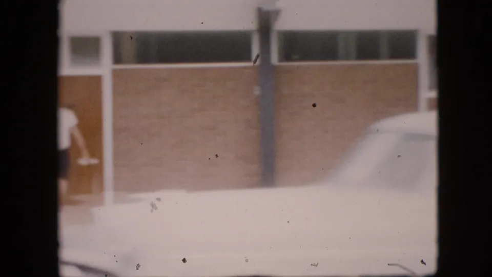 In 1958 Pennsylvania home-movie footage, a partial man in a white shirt and dark shorts stands near a glass-panel doorway beside a brick building and a white car.