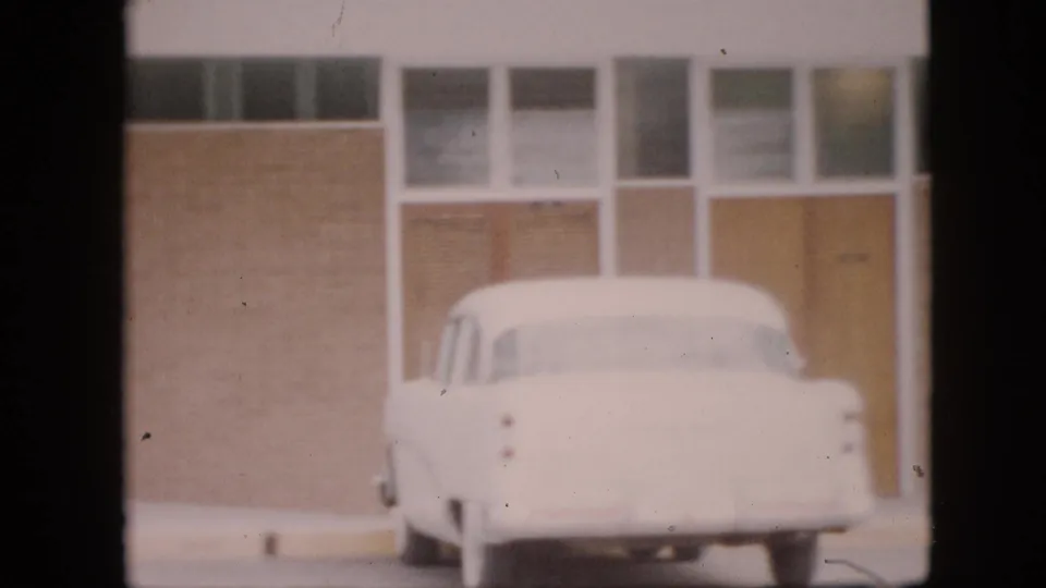 A white mid-century car is parked outside a brick building with rectangular glass panels and doors in the Pennsylvania 1958 home-movie scene.