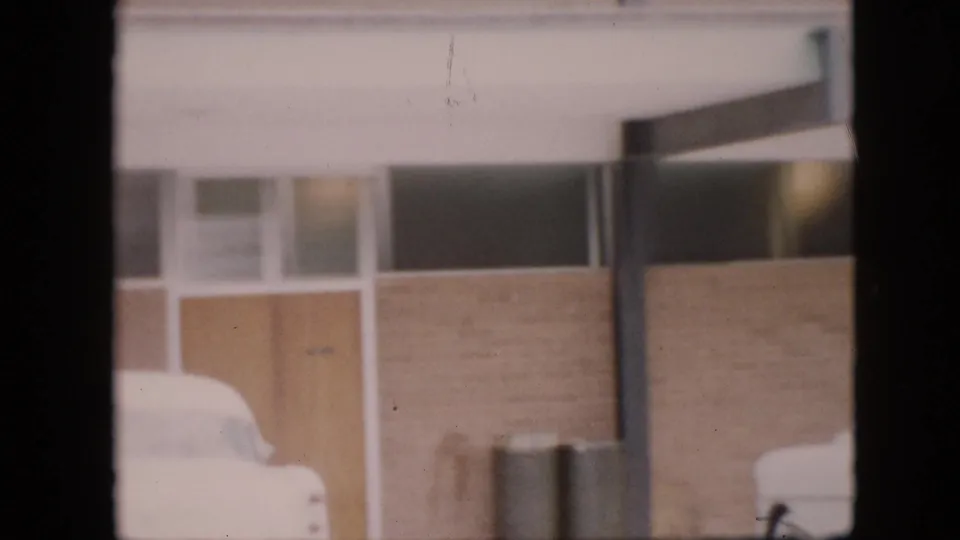 The camera frames the brick building entrance, flat canopy, glass-panel doors, and a partial white car during the 1958 Pennsylvania exterior shot.
